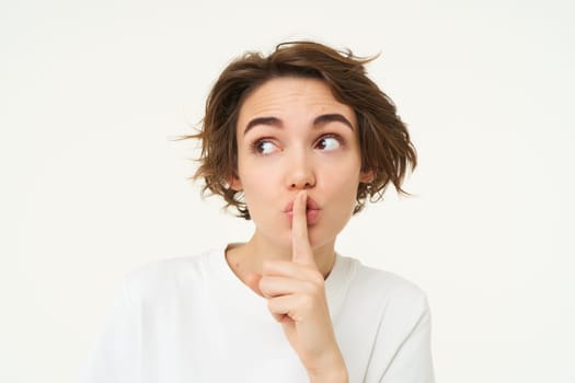 Cute and coquettish woman, keeps secret, gossips, holds finger on lips, shushing, hush gesture, stands over white background.