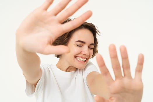 Close up of happy brunette woman extending her hands, stretching, covering herself from camera, laughing and smiling, standing over white background.