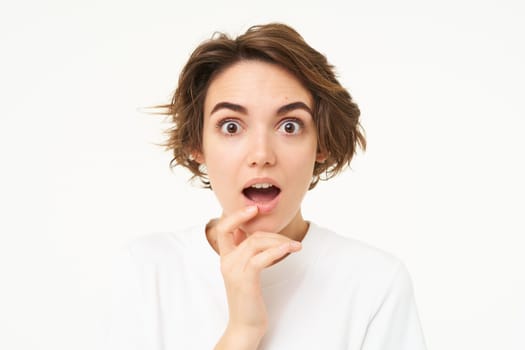 Close up of amused, intrigued young woman, drops jaw and listens with interest, stands over white background.