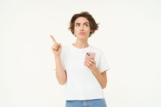 Portrait of woman thinking, using smartphone and looking thoughtful, making choice, deciding, standing isolated over white background.