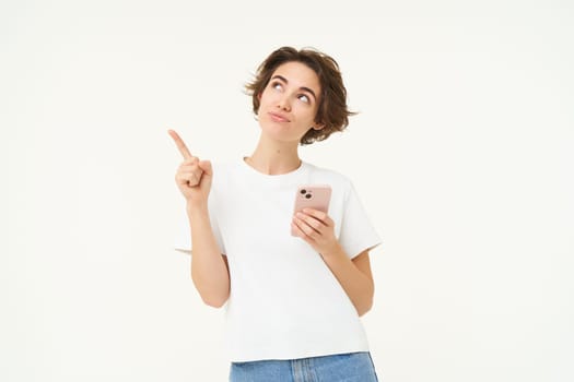 Portrait of young modern woman with mobile phone, looking up and pointing at advertisement, standing against white studio background.