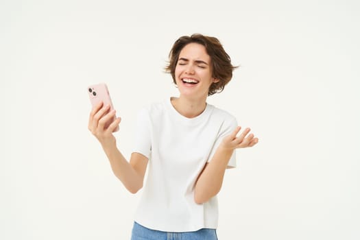 Portrait of chatty young woman, laughing, using mobile phone, video chats online on smartphone, isolated over white background.