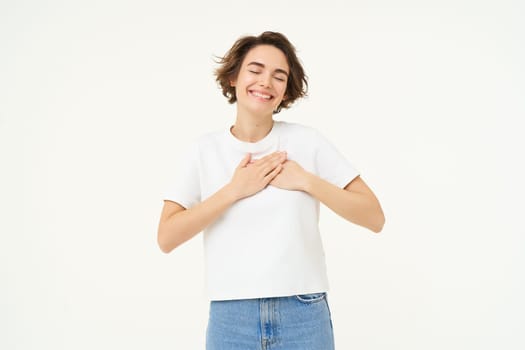 Image of brunette woman holding hands on heart, express gratitude, heart-warming feelings, emotion of happiness, love and care, isolated over white background.