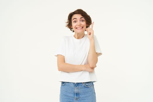 Excited girl has an idea, suggests a plan, points up, raises one index finger and smiles, has solution, stands over white background.