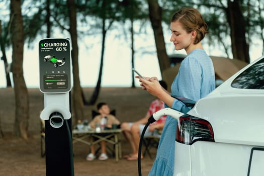 Outdoor adventure and family vacation camping at sea travel by eco friendly car. Woman or mother check car's battery with smartphone while charging EV car frin charging station in campsite. Perpetual