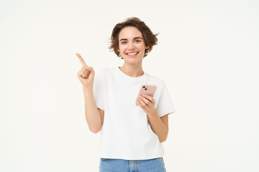 Portrait of smiling, carefree woman, holding smartphone, pointing finger at copy space, advertisement, using mobile phone and recommending store, standing over white background.