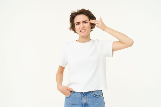 Portrait of woman holding fingers near temple, beeing fed up, annoyed by something, standing tired against white background.