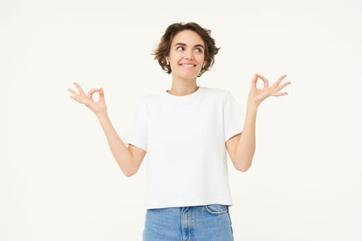 Portrait of young girl feeling happy and positive, meditating, holding hands sideways in zen, practice yoga, standing over white background. Copy space