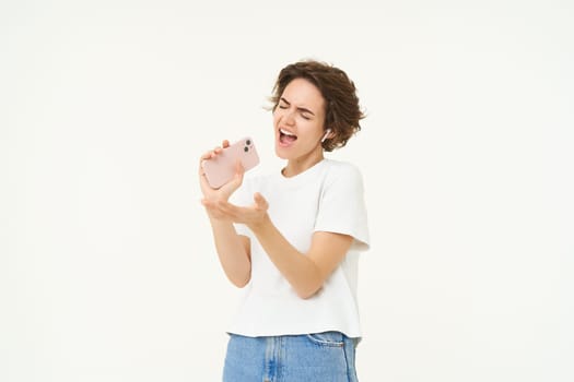 Portrait of happy young woman singing in mobile speakerphone, listening music in wireless headphonse, playing smartphone karaoke game, standing over white background.