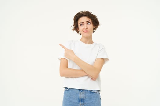 Image of woman with sceptical expression, pointing finger left, showing advertisement with doubtful face expression, standing over white studio background.