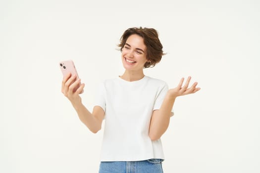 Potrait of woman video chats with friend, showing around, demonstrating something, has an online meeting on smartphone, standing over white background.