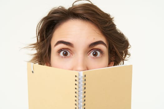 Close up portrait of girl face, hiding behind notebook, looking aside, standing over white background.