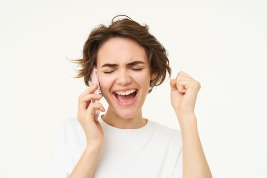 Excited brunette woman celebrating, answers phone call and looking happy, winning, triumphing from great big news, standing over white background.