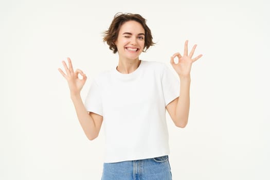 Cheerful brunette woman shows okay, ok sign and smiling, gives her approval, recommanding, saying yes, zero problem, standing over white background.