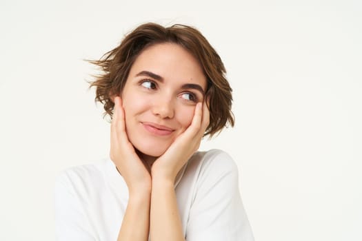 Close up of hopeful young woman, girl anticipating something, touching her face, looking forward to something, waiting with excitement, standing over white background.