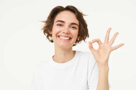 Close up portrait of satisfied, smiling young woman, shows okay, ok gesture, recommends something, gives positive feedback, says yes, white background.