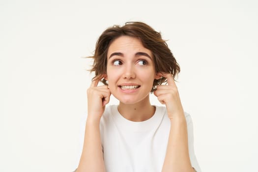 Close up portrait of woman smiling with awkward face expression, shuts her ears, doesnt want to listen, disturbed by loud noise, stands over white studio background. Copy space