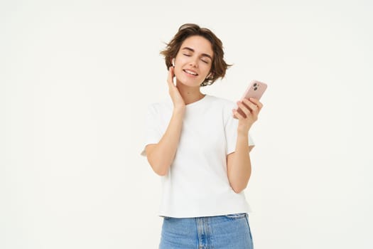 Portrait of smiling, beautiful young woman in wireless headphones, puts on music in earphones, holds smartphone, using streaming app, standing over white background.