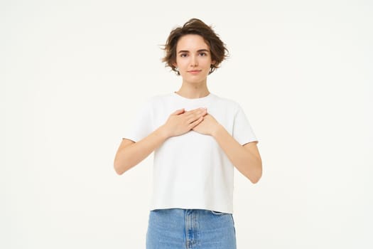Portrait of brunette woman with hands on chest, looks with care and love, express gratitude, self-care and heart-warming feelings, stands over white background.