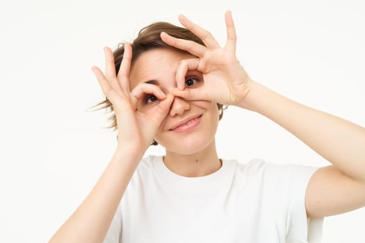 Close up of carefree, cute young woman showes finger glasses over her eyes and smiling, isolated over white background. Copy space