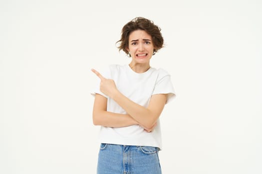 Portrait of woman feels discomfort, points finger left, shows advertisement and looks concerned, stands over white studio background.