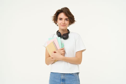 Image of brunette woman in headphones, standing with documents. Student with notes and workbooks posing against white background.