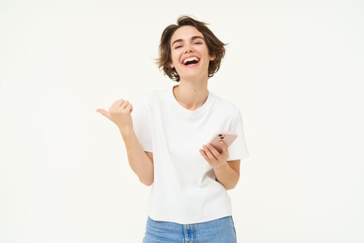 Portrait of smiling, carefree woman, holding smartphone, pointing finger at copy space, advertisement, using mobile phone and recommending store, standing over white background.