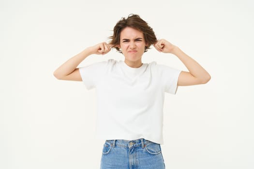 Portrait of woman disturbed by loud sound, shuts her ears, annoyed by music, frowning, standing over white background.