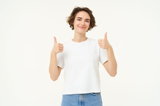 Image of young woman giving positive feedback, shows thumbs up and smiling, looks with confidence, stands over white background. Copy space