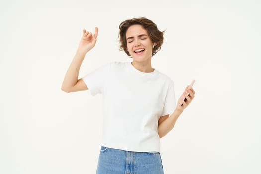 Portrait of woman singing and listening music in wireless headphones, holding mobile phone, having fun, posing over white studio background.