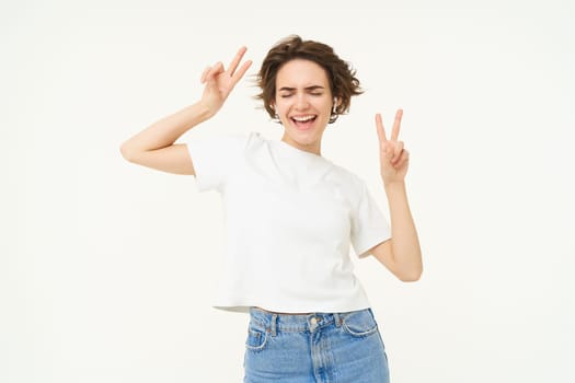 Carefree woman dancing, listening to music in wireless headphones, dancing and showing peace sign, white studio background.