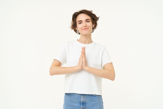 Portrait of mindful young woman, showing namaste, grateful gesture, saying thank you, asking for something and smiling, standing over white background.