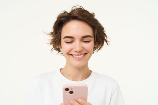 Close up portrait of young woman smiling, using smartphone, texting, checking her mobile apps, stands over white background. Copy space