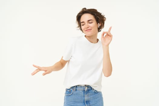 Portrait of carefree, happy young woman in wireless headphones, listens to music, dances to favourite song in earphones, posing over white studio background.