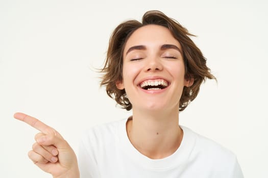 Close up portrait of brunette girl with happy face, laughing, pointing finger left, showing advertisement, standing over white studio background. Copy space