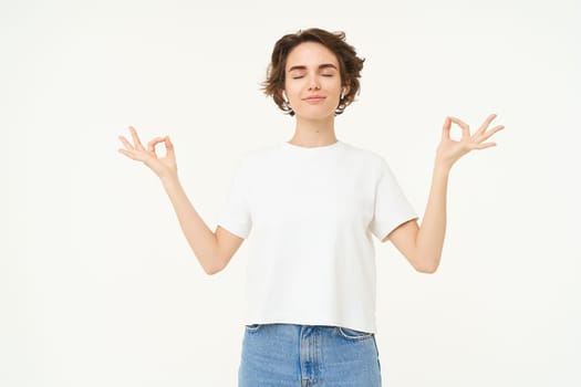Portrait of young woman feeling peace and relaxation, holding hands sideways, zen gesture, meditating, practice mindfulness yoga, standing over white background.