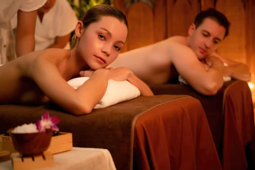 Caucasian couple customer enjoying relaxing anti-stress spa massage and pampering with beauty skin recreation leisure in warm candle lighting ambient salon spa at luxury resort or hotel. Quiescent