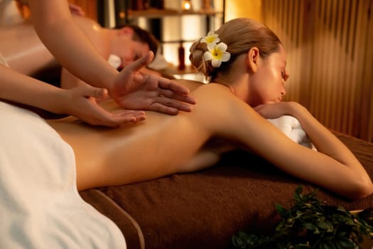 Caucasian couple customer enjoying relaxing anti-stress spa massage and pampering with beauty skin recreation leisure in warm candle lighting ambient salon spa at luxury resort or hotel. Quiescent