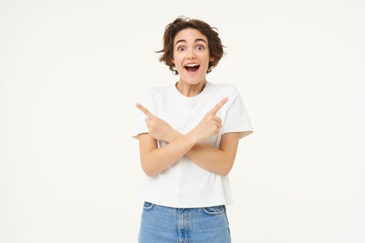 Portrait of amazed, happy young woman pointing sideways, left and right direction, showing advertisement, two choices, standing over white studio background.