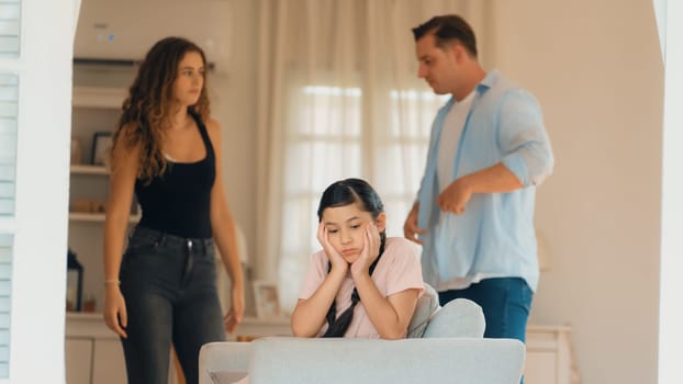 Annoyed and unhappy young girl sitting on sofa trapped in middle of tension by her parent argument in living room. Unhealthy domestic lifestyle and traumatic childhood develop to depression.Synchronos