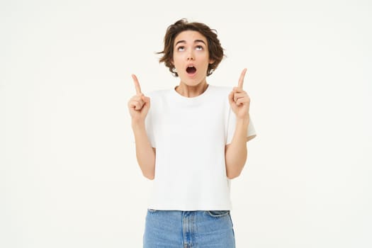 Portrait of woman with surprised face, amazed by promo offer, pointing fingers up and looking on top, standing against white background.