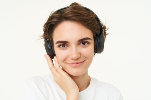 Image of young modern woman in wireless headphones, using earphones to listen to music, standing over white background. Copy space
