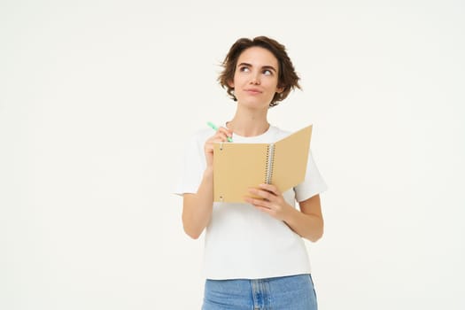 Portrait of creative girl, student doing homework, writing in notebook, standing with her diary, posing over white background.