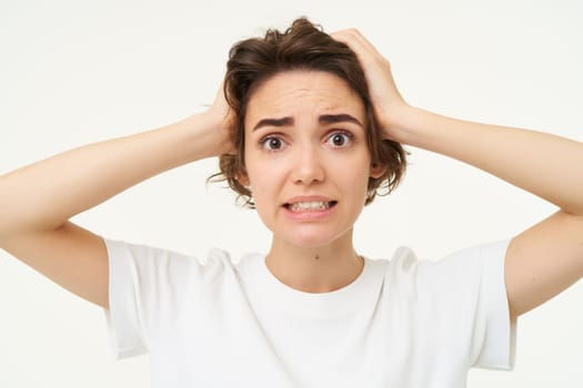 Close up of woman in panic, holding hands on head and clenching teeth, looking troubled, puzzled and frustrated by something, standing over white background.