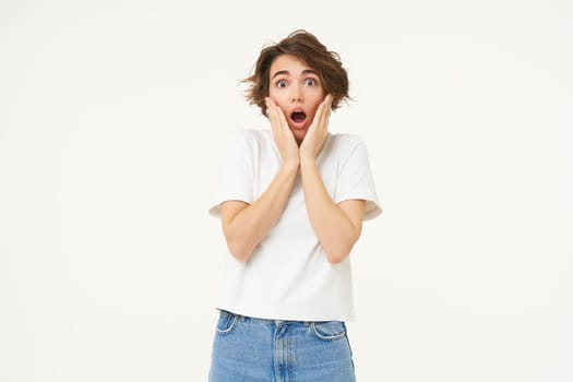 Portrait of girl with shocked, scared face, screams, holds hands on cheeks, stands over white studio background.