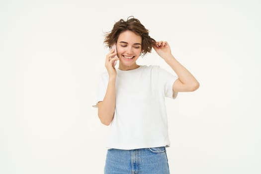 Cute and coquettish young woman calling someone, answer telephone, talking on smartphone and playing with her hair, standing over white background.