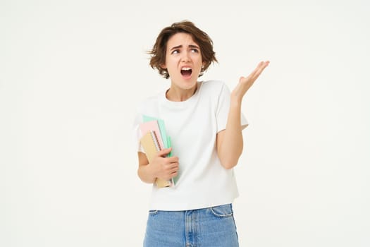 Image of shocked young woman, holding notebooks, shrugging and looking disappointed, complaining at something and staring at upper right corner advertisement, white background.