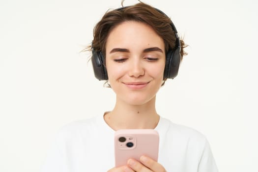 Portrait of candid young woman, listens music in wireless headphones, holding smartphone, watching videos on mobile phone, standing over white background.