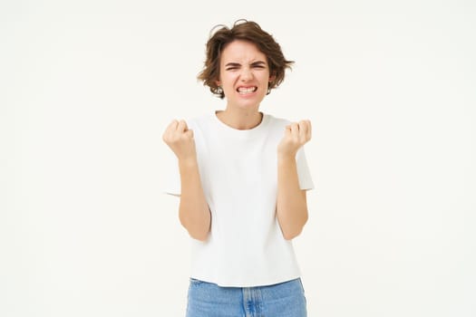 Portrait of confident and motivated woman, clenches her fists and looks self-assured, stands over white background. Copy space