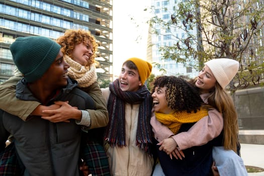Happy multiracial young friends enjoying winter day outdoors. Cheerful group of smiling friends piggyback ride outdoors in the city. Lifestyle concept.